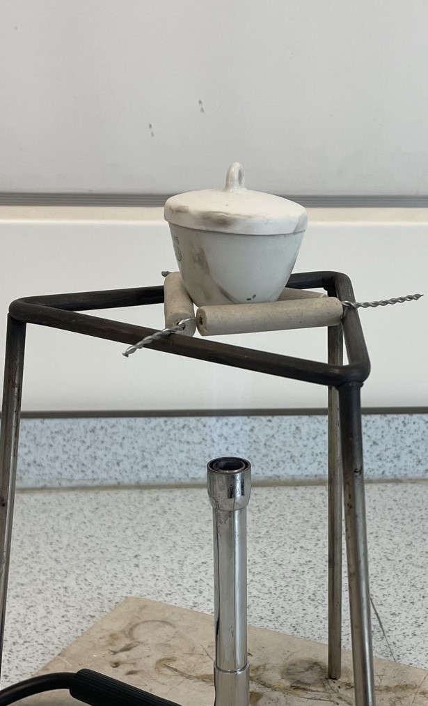 a photograph of a crucible sitting in a clay triangle balanced on a tripod above a Bunsen burner